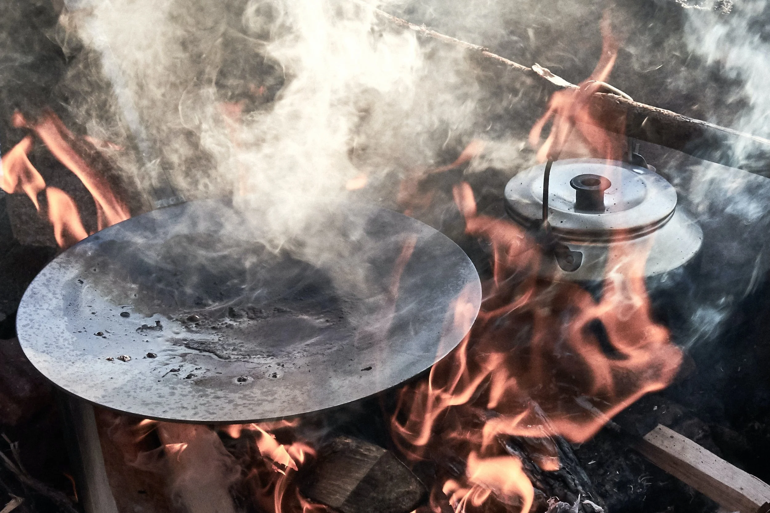 A metal plate and a kettle are heating over an open fire with visible flames and smoke.