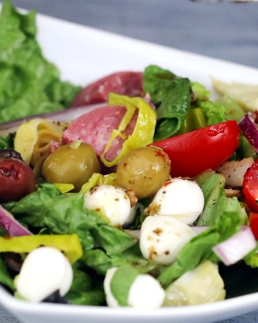 Fresh vegetable salad with lettuce, tomatoes, olives, onions, mozzarella, and sliced meats in white bowl