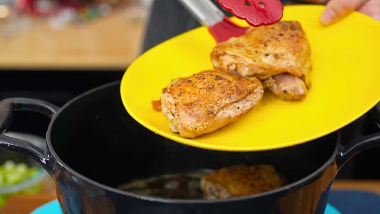 Golden-browned chicken thighs lifted from a Dutch oven onto a yellow plate with red tongs