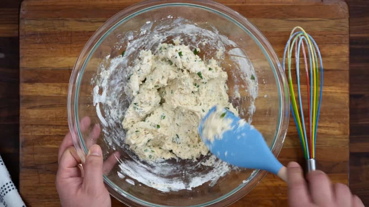 Overhead shot of fluffy dumpling dough flecked with parsley being mixed in a glass bowl with a blue spatula