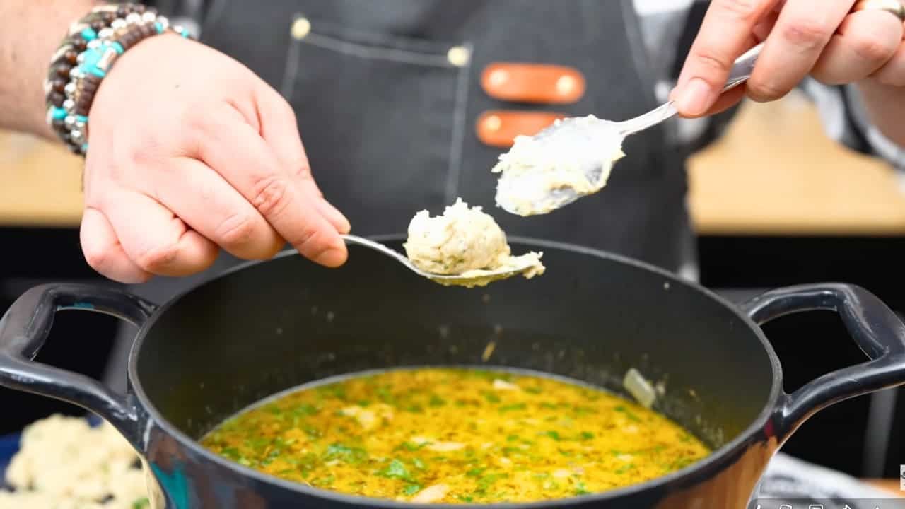 A spoonful of dumpling dough being dropped into a pot of simmering herbed chicken broth