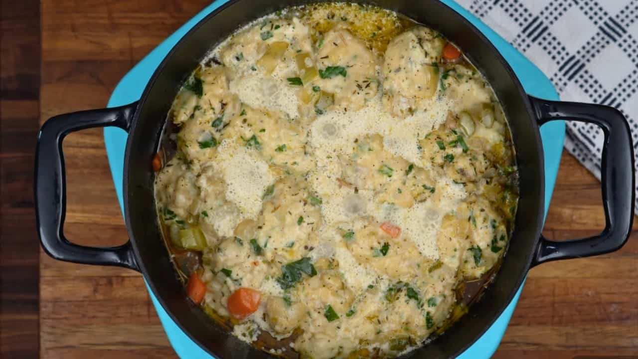 Overhead view of finished creamy chicken and dumplings with carrots, peas, and fresh herbs in a black Dutch oven