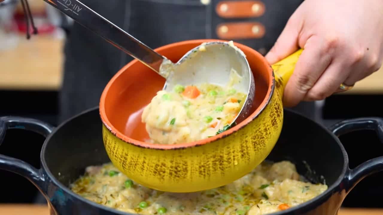 Ladling creamy chicken and dumplings with peas and carrots into a yellow-and-orange ceramic bowl