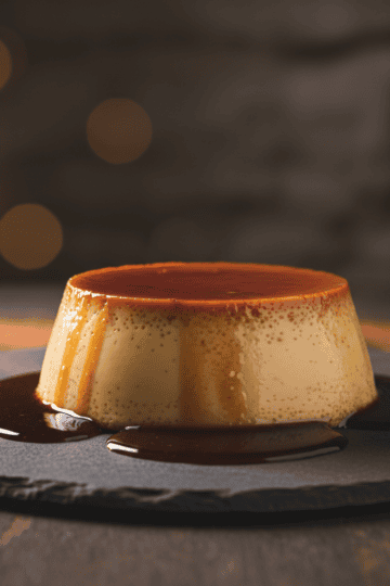Dominican flan de leche unmolded on dark slate with dark amber caramel sauce pooling around the base