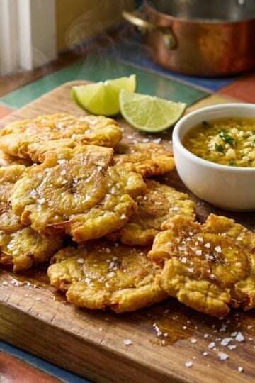 Golden crispy Dominican tostones piled on a wooden board with mojo de ajo garlic sauce, lime wedges, and cilantro