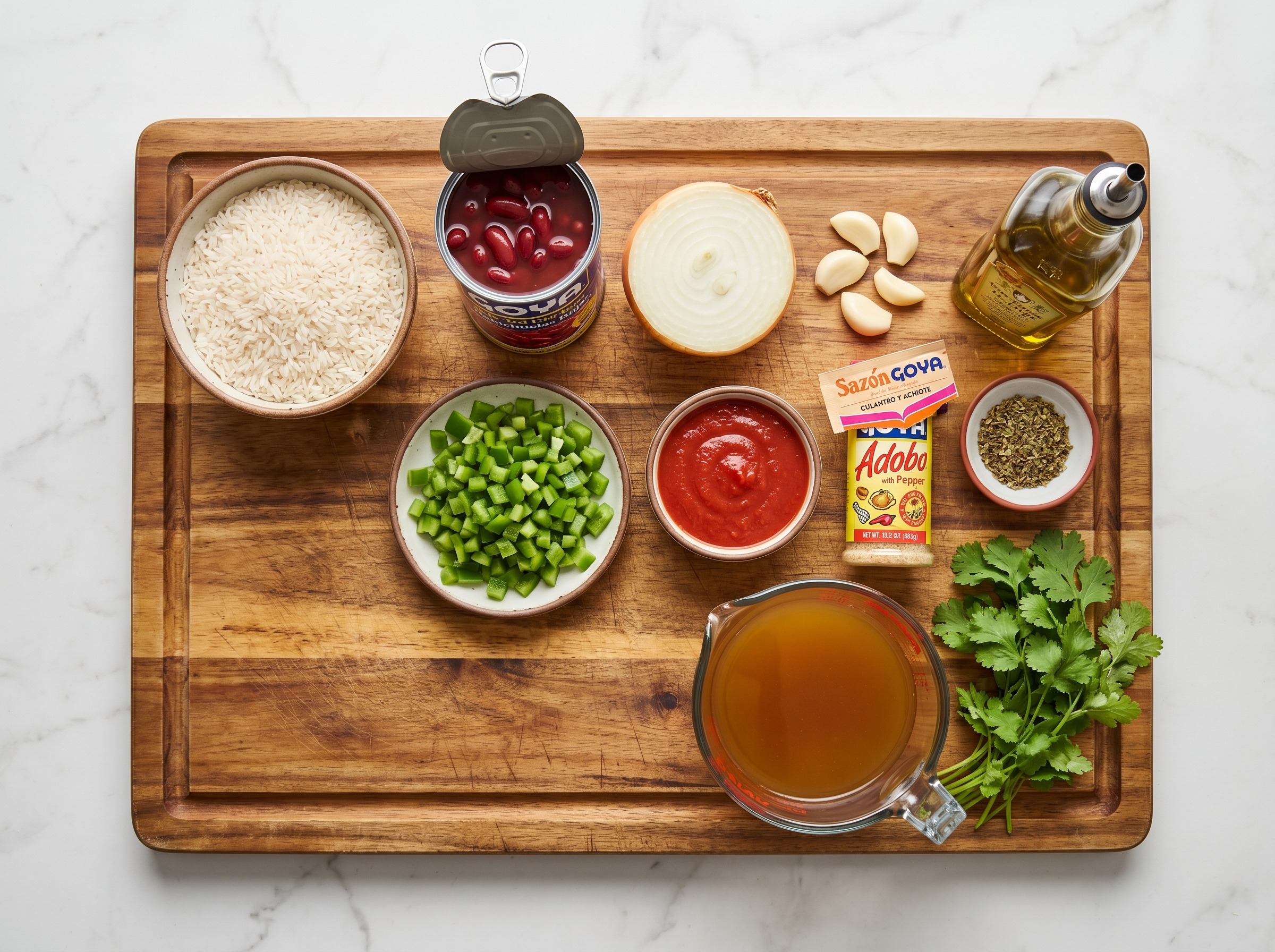 All ingredients for moro de habichuelas rojas laid out on a wooden cutting board: rice, canned red kidney beans, onion, garlic, bell pepper, tomato sauce, sazón, adobo, oregano, olive oil, cilantro, and chicken stock