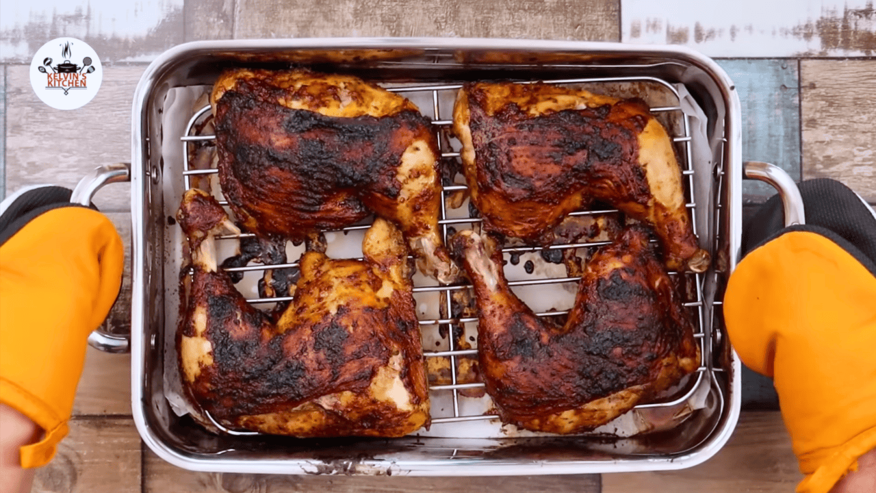 Four golden brown Peruvian chicken leg quarters on a roasting rack in a stainless steel pan