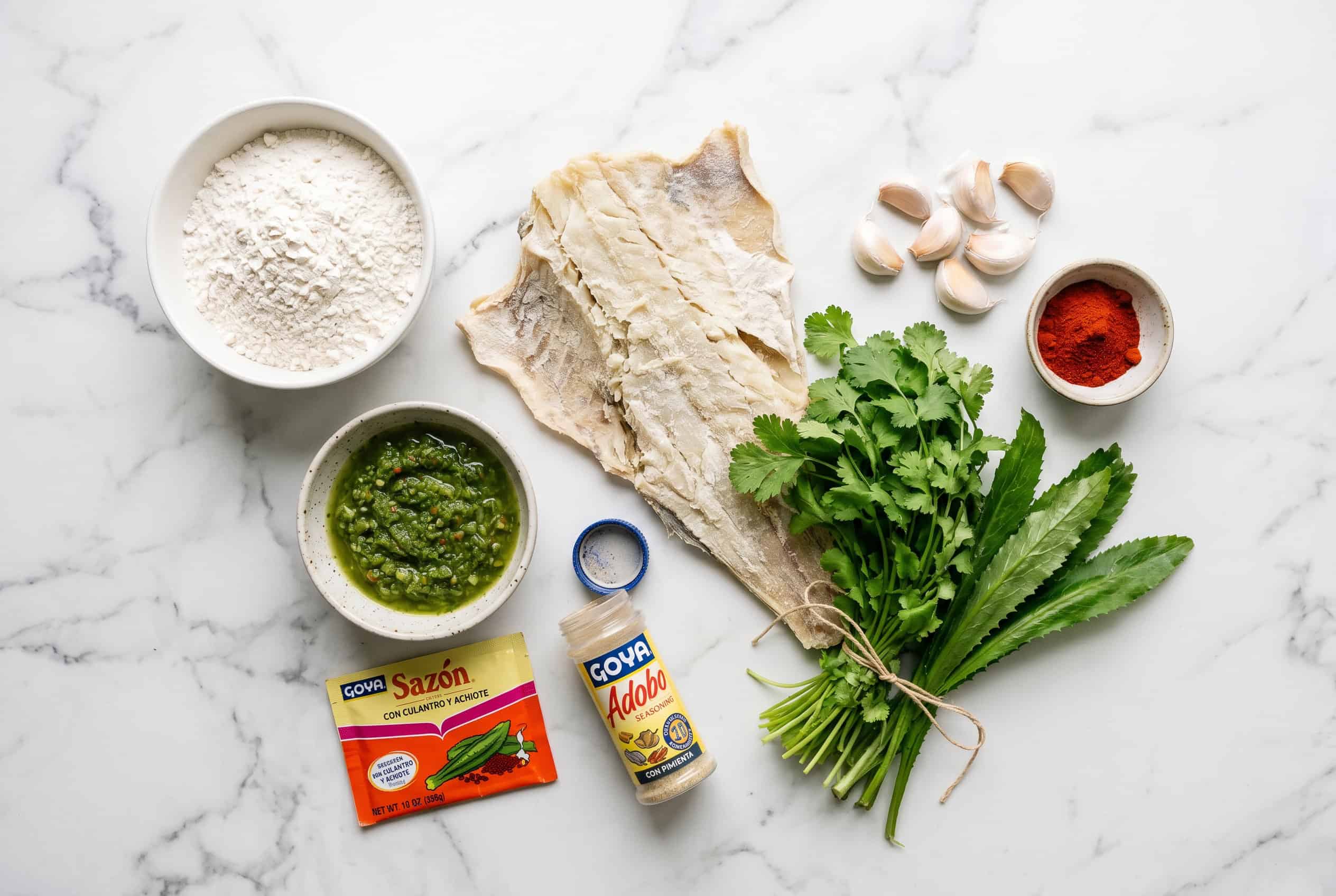 Overhead flat lay of Puerto Rican bacalaitos ingredients: salt cod, flour, sofrito, adobo, sazón, garlic, cilantro