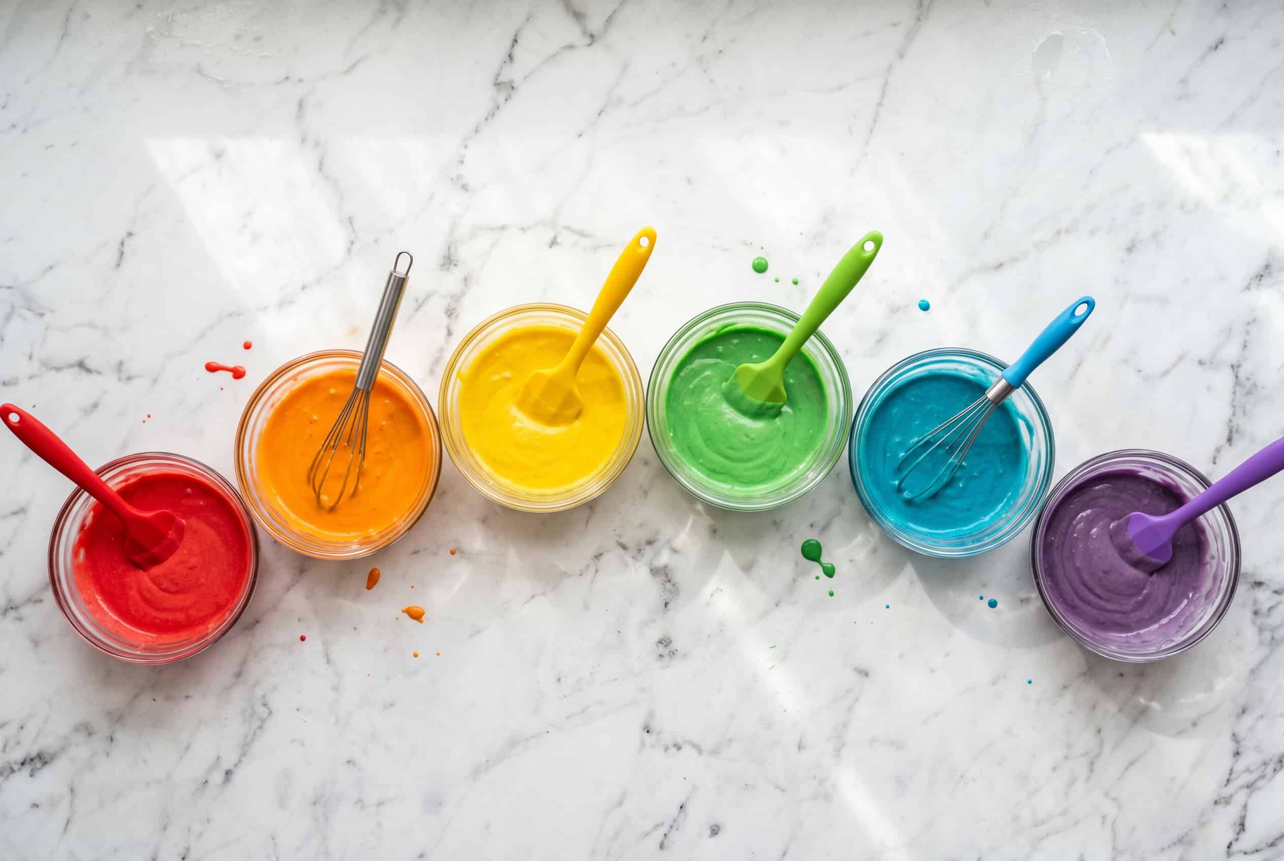 Six small glass bowls of rainbow-colored cake batter arranged on a white marble countertop, overhead view