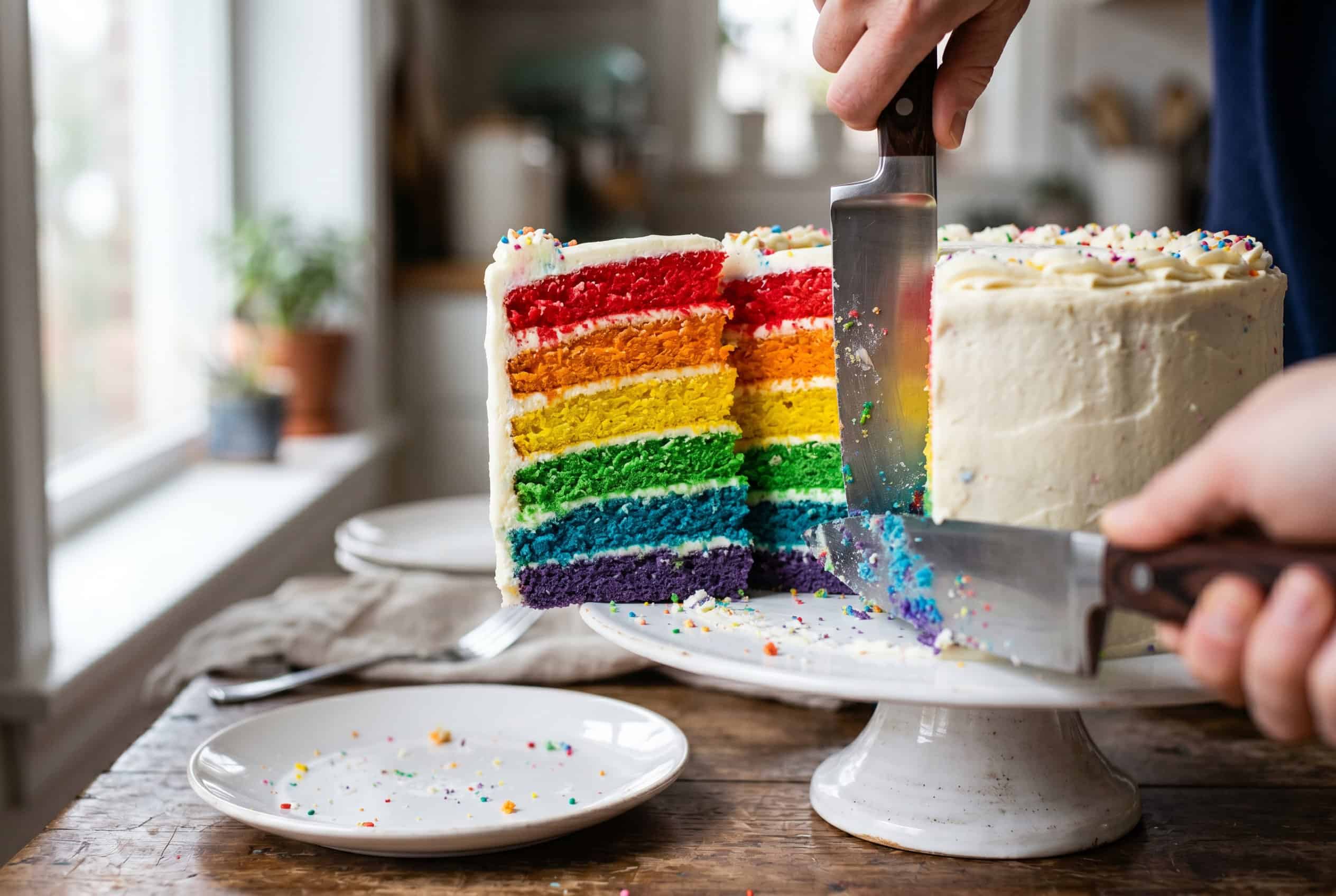 A sharp kitchen knife slicing through a frosted rainbow layer cake, revealing the vivid red, orange, yellow, green, blue, and violet layers inside