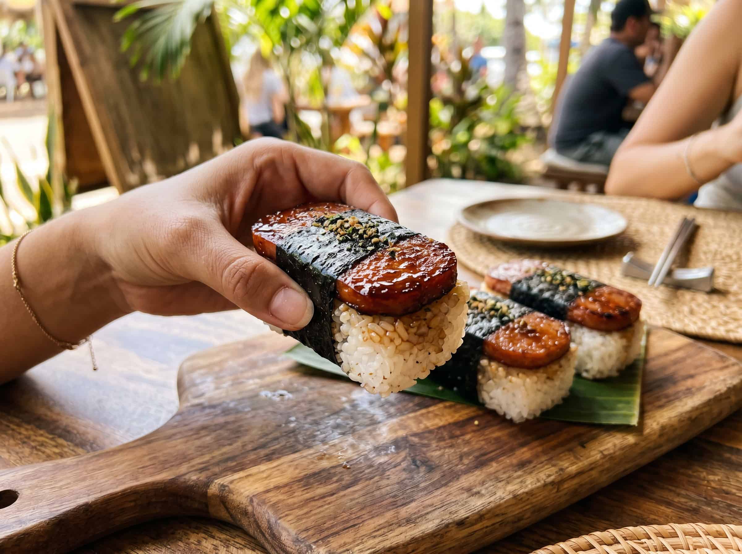 A hand serving a piece of Hawaiian spam musubi from a wooden board with more musubi pieces in the background