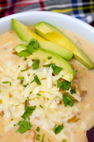Creamy slow cooker white chicken chili in a ceramic bowl topped with avocado slices, shredded cheese, and a blue corn tortilla chip