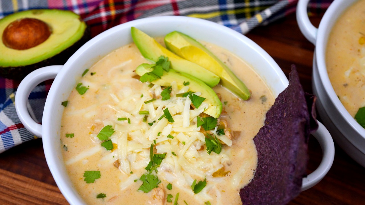 Creamy slow cooker white chicken chili in a ceramic bowl topped with avocado slices, shredded cheese, and a blue corn tortilla chip