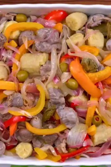 Overhead view of Caribbean chicken gizzard escabeche in a white casserole with green bananas, peppers, onions, and olives