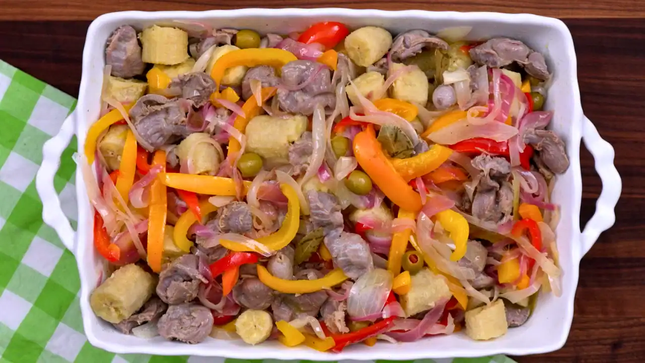 Overhead view of Caribbean chicken gizzard escabeche in a white casserole with green bananas, peppers, onions, and olives