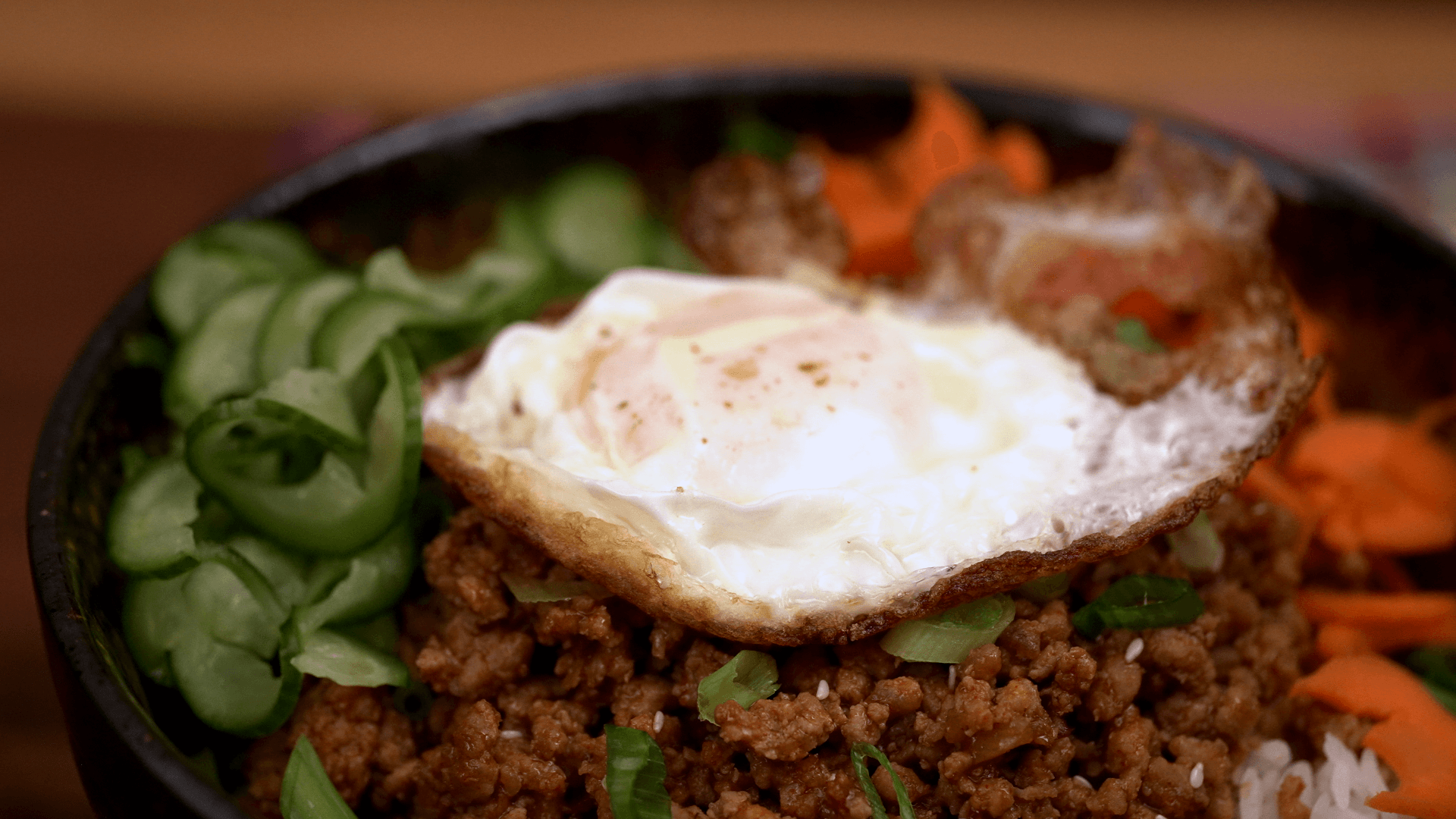 Close-up of gochujang pork bowl with crispy fried egg, cucumber ribbons, and shaved carrots — Korean rice bowl recipe