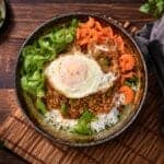 Gochujang pork bowl with fried egg, sliced cucumbers, shaved carrots, and scallions over rice on a bamboo mat