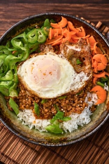 Gochujang pork bowl with fried egg, sliced cucumbers, shaved carrots, and scallions over rice on a bamboo mat
