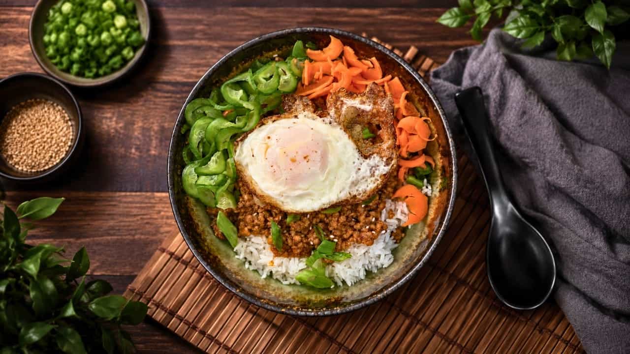 Gochujang pork bowl with fried egg, sliced cucumbers, shaved carrots, and scallions over rice on a bamboo mat