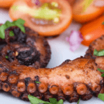 Best Grilled Octopus Recipe Grilled octopus tentacles, charred and seasoned, garnished with fresh parsley, with cherry tomatoes and red onion on a white plate