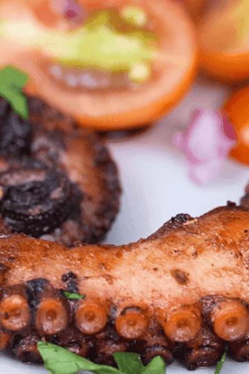 Grilled octopus tentacles, charred and seasoned, garnished with fresh parsley, with cherry tomatoes and red onion on a white plate