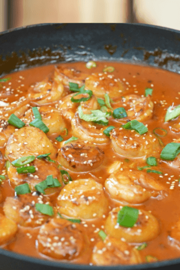 Spicy gochujang scallops and shrimp simmering in a creamy red sauce, topped with sesame seeds and green onion, in a skillet